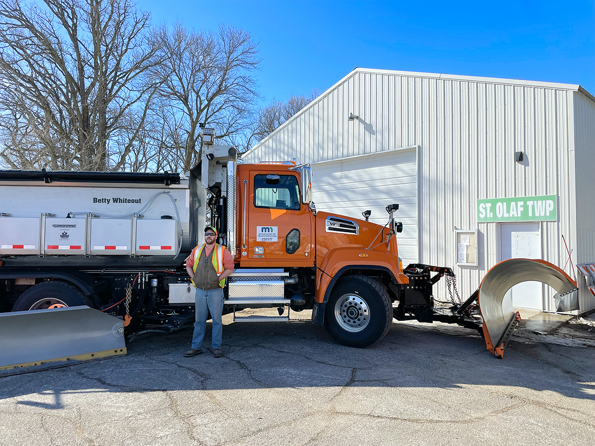 PHOTO: Snowplow driver standing alongside orange snowplow truck with the name, Betty Whiteout, on its side. Truck is next to building that says "St Olaf Twp"